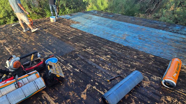 Two people work on a weathered, flat rooftop partially covered with blue sealant. Tools, rollers, and materials are scattered around, with trees and greenery visible in the background.