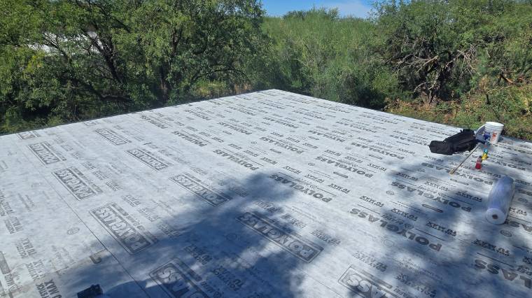 Roof covered with a layer of roofing underlayment labeled “POLYGLASS,” surrounded by trees and greenery under a clear blue sky. Roofing tools and materials are visible on the right side.