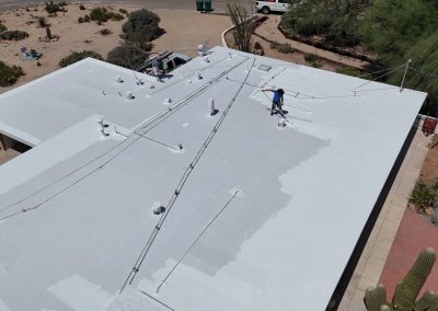 A person uses a spray machine to apply a white coating on a flat rooftop of a building surrounded by desert landscaping, with equipment and cables visible on the roof.