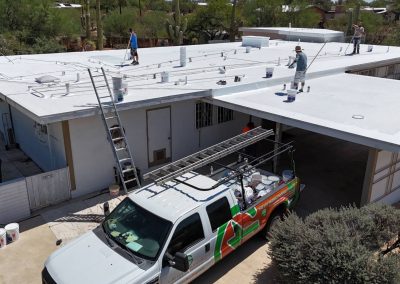 Two workers are applying white coating to the flat roof of a large building. A utility truck with a ladder on top is parked below, and paint buckets are placed on the roof and ground. Desert vegetation surrounds the area.