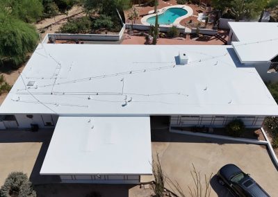 Aerial view of a flat-roofed white house with surrounding driveway, desert landscaping, and a backyard featuring a swimming pool and patio area. A black car is parked in the driveway.