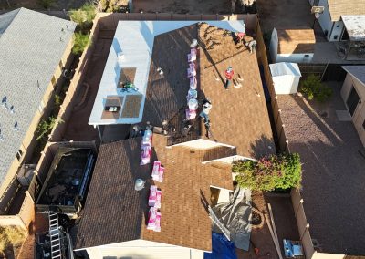 Aerial view of workers installing new shingles on the brown roof of a house; materials are stacked along the roof, and equipment is visible in the yard below. Neighboring houses are seen nearby.