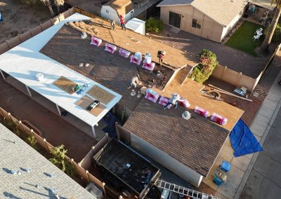 Aerial view of workers installing new roofing materials on a house. Roofing supplies are spaced along the roof, some sections are covered with white sheeting, and tools are scattered. Adjacent houses, a street, and vehicles are visible.