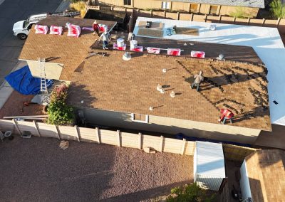 Aerial view of workers replacing shingles on a brown residential roof; bundles of roofing materials are stacked on the roof, and some workers are installing new shingles. There is a white roof section and fencing around the yard.