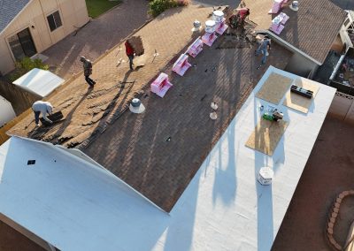 Aerial view of workers repairing the roof of a house. One side of the roof is stripped of shingles, while the other side has workers, roofing materials, and tools. The yard and nearby buildings are visible.