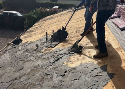 Two workers are removing old, damaged roofing shingles from a rooftop with roofing tools. Pieces of shingles are scattered across the exposed wooden roof surface. Sunlight casts shadows on the work area.