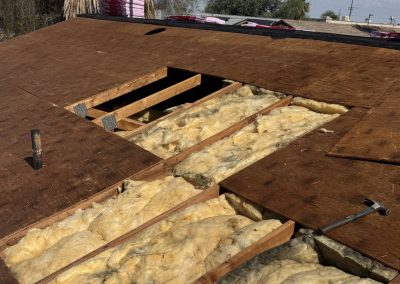 A roof under repair with several plywood boards removed, exposing yellow insulation and wooden beams underneath. Tools, including a hammer, are lying on the roof.