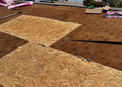 A section of a roof with sheets of plywood, some new and lighter in color, fitted among older, darker boards. Roofing materials and tools are scattered nearby, indicating roof repairs in progress.