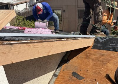 Two workers on a sloped roof are installing insulation and roofing materials. One person kneels with pink insulation, while the other uses tools near the roof edge. Exposed wood and roofing layers are visible in the foreground.