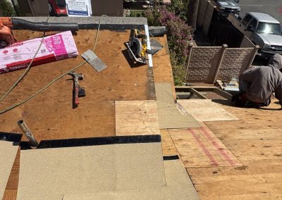 A person in a gray hoodie works on a sloped wooden roof under construction, surrounded by tools, insulation boards, and roofing materials on a sunny day. Vehicles and houses are visible in the background.