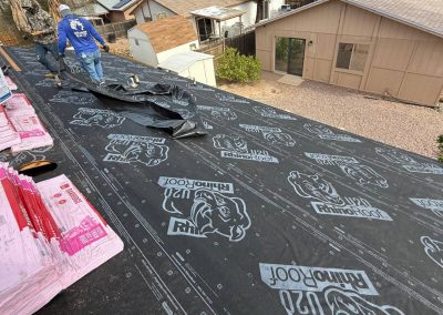 Two workers install roofing underlayment labeled "RhinoRoof" on a house roof, with shingles and supplies nearby. Several houses and a yard are visible in the background.