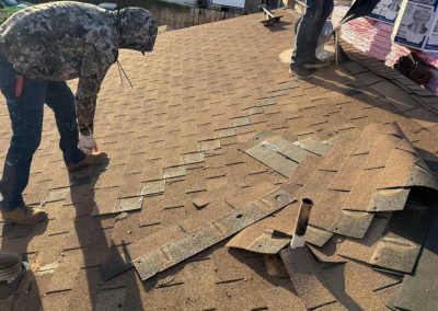 A person in a camouflage hoodie and boots repairs brown roof shingles, while roofing materials and tools are scattered nearby on the sloped rooftop in sunlight.