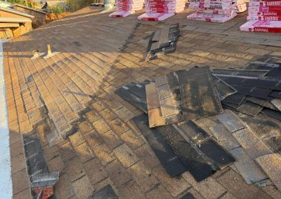 A house roof with several stacks of new shingles and partially installed brown shingles. Some old shingles are removed, and tools are visible on the roof, indicating ongoing roofing work.