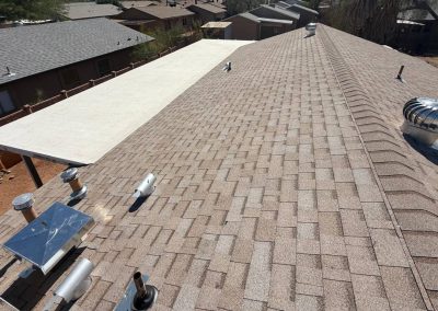 A view of a residential roof with brown asphalt shingles, metal vents, and pipes. Adjacent houses and a covered patio area are visible in the background under a clear sky.