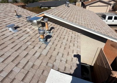 Rooftop view of a house with light brown shingles, several vent pipes, and metal fixtures. Nearby houses and a white truck are visible in the background on a sunny day.