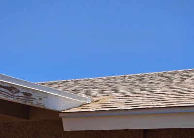 A close-up of a house roof with brown shingles, a rusted metal vent pipe, and peeling paint on the wooden eaves, under a clear blue sky.