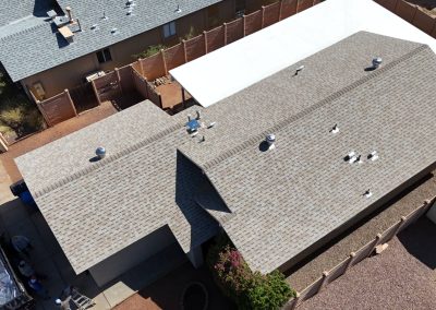 Aerial view of a residential house with a gray shingle roof, white roof on the neighboring building, and a fenced backyard with gravel landscaping and a small shed.