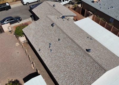 Aerial view of a residential house with a light gray shingle roof, surrounded by a gravel yard, neighboring houses, and parked cars along a street.
