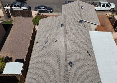 Aerial view of a house with a shingle roof, a gravel yard to the left, and vehicles parked along the street and driveway. The roof has several vents and skylights. Neighboring buildings are visible on either side.