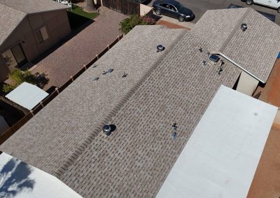 Aerial view of a residential roof with gray shingles, several roof vents, and a section of flat white roofing; cars and landscaped yards are visible nearby.