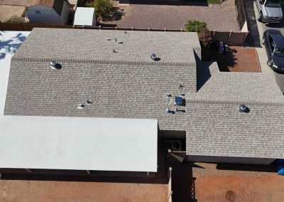 Aerial view of a house with a gray shingled roof, white carport on the left, and a driveway on the right leading to a street with parked cars. Some backyard and surrounding fence are also visible.
