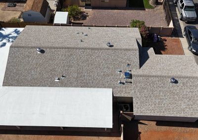 Aerial view of a house with a gray shingle roof and a white flat roof extension. The yard has a mix of dirt, gravel, and paved areas. Several cars are parked along the street and driveway.