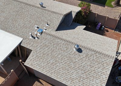 Aerial view of a house with a gray shingle roof, multiple roof vents, and a small covered porch. Part of the driveway, parked cars, and a group of people are visible at the edge of the image.