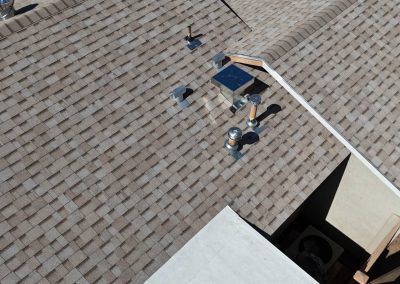 Aerial view of a beige shingle roof with several vent pipes, metal vents, and a turbine vent. An open section reveals part of the structure beneath the roof.