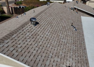 A view of a residential roof with asphalt shingles, several vent pipes, and a metal turbine vent. The roof is part of a single-story house, with neighboring homes and vehicles partially visible.