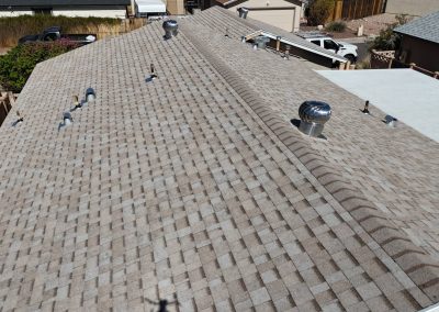 Aerial view of a house roof with light brown asphalt shingles, several vent pipes, and a silver turbine vent, surrounded by a fenced yard and other nearby houses.