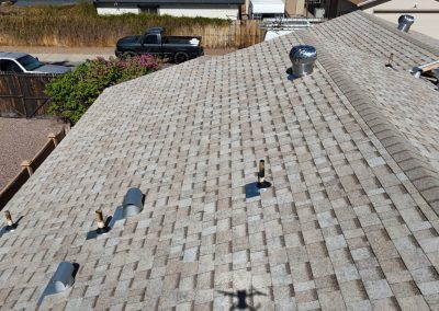 Aerial view of a house roof with light gray asphalt shingles, several roof vents, pipes, and a turbine vent; a yard, fence, and parked vehicles are visible in the background.