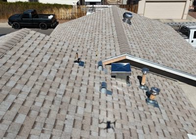 Aerial view of a house’s shingled roof with several vent pipes and a metal vent. Two pickup trucks are parked on the street below, and there is a shadow of the drone on the roof.