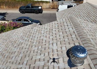 A rooftop with patterned shingles, a silver roof vent, and the shadow of a drone. Below, two parked vehicles—a black pickup truck and a dark sedan—are on the street next to a white van and a wooden fence.