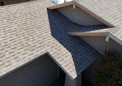 A close-up view of a house roof with light brown asphalt shingles, intersecting rooflines, and part of a flowering bush visible at the lower right corner.