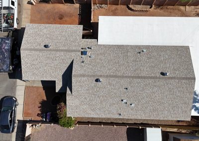 Aerial view of a house with a gray shingle roof, a white flat-roof extension, two parked vehicles on the street, and a fenced backyard with some landscaping and a tree casting a shadow on the roof.