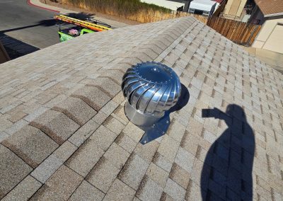 A silver roof turbine vent is installed on a gray shingle roof in sunlight. A person’s shadow holding a phone is visible on the roof, as well as a residential street and other houses in the background.