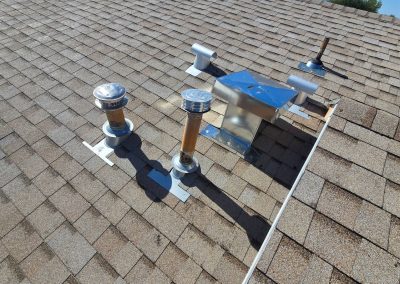 Tan shingle roof with several metal vent pipes and a rectangular metal box, likely part of the HVAC or plumbing system, securely mounted and casting shadows in bright sunlight.