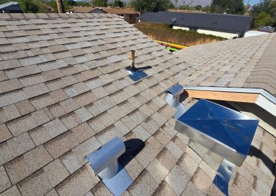A rooftop with brown shingles, several shiny metal vents, a pipe, and a rectangular metal structure. Some exposed wood is visible near the roof’s peak, with houses and trees in the background under a clear sky.