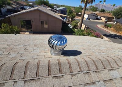A shiny metal roof vent sits on a shingled rooftop of a house in a suburban neighborhood, with houses, trees, and mountains visible in the background under a clear blue sky.