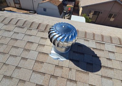A shiny metal roof turbine vent is installed on a shingled roof, casting a distinct shadow. Nearby houses with similar roofs are visible in the background under bright sunlight.