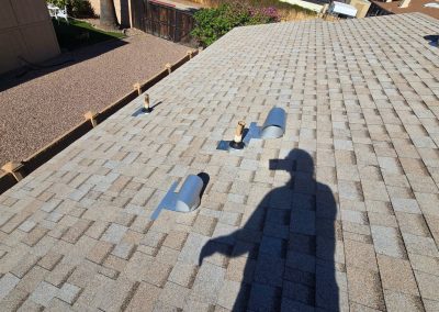 Shadow of a person on a sloped shingle roof with multiple vents and pipes, next to a gravel yard and a building in the background, under bright sunlight.