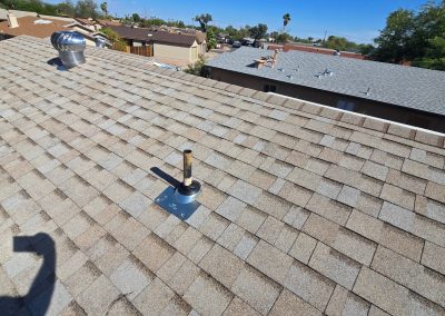 A rooftop with light brown shingles, a metal vent pipe, and a turbine vent. Neighboring roofs and trees are visible in the background under a clear blue sky. A person's shadow is visible on the roof.