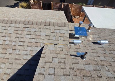 A rooftop with brown and gray asphalt shingles, several metal vents, and pipes. A small gap is visible where two roof sections meet. There are wooden fences and a white canopy in the background.