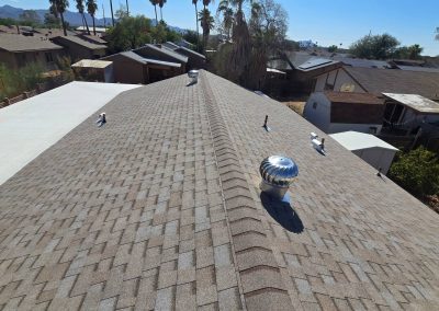 A rooftop view of a residential neighborhood shows gray asphalt shingles, metal roof vents, and nearby houses with similar roofs. Palm trees and clear blue skies are visible in the background.