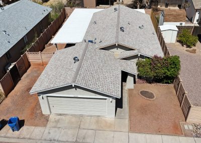 Aerial view of a single-story house with a light gray roof, attached garage, and minimal landscaping. The front yard has bare dirt, a small bush, and is bordered by neighboring homes and wooden fences.