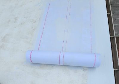 A rolled-up sheet of white waterproof membrane is partially unrolled on a flat, white rooftop next to a brick pavement and a gray stone wall.