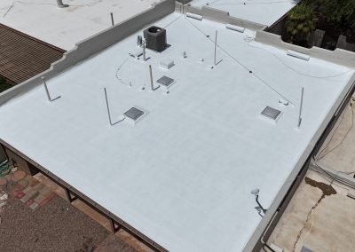 Aerial view of a flat, white-coated rooftop with several vents, pipes, skylights, and an HVAC unit. The surrounding area includes patios, landscaping, and neighboring buildings.