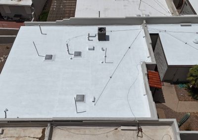 Aerial view of a flat white roof on a building with multiple vents, skylights, and a central HVAC unit. The surrounding area includes neighboring roofs, a small garden, and paved pathways.