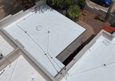 Aerial view of white flat rooftops connected by cables, with a round vent on one roof, a blue car parked beside a desert garden, and various cacti and shrubs visible in the scene.