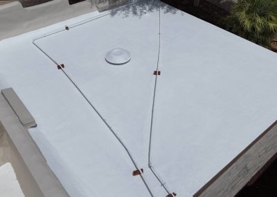 A flat white roof with a round skylight, white pipes secured by brown brackets, and a small palm tree visible nearby. The roof is bordered by light-colored walls and a yard.